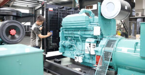 A technician in a factory workshop inspects a large, light blue industrial diesel generator set.